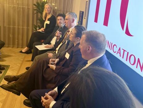 Picture of panel sat on stage in front of big screen with giant red logo on it, one man is holding microphone and talking