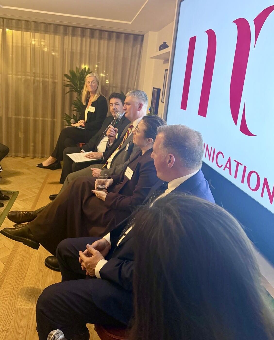 Picture of panel sat on stage in front of big screen with giant red logo on it, one man is holding microphone and talking