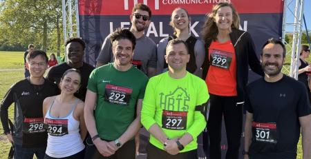 Group of 9 men and women posing for photo in park in front of backdrop that reads MD Communications Legal 5k run. Two women and a man are standing on podium. All wear running clothes and have race numbers pinned to them.