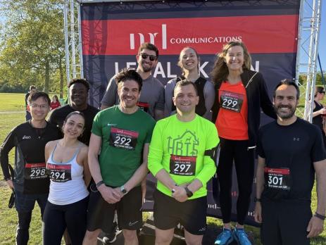 Group of 9 men and women posing for photo in park in front of backdrop that reads MD Communications Legal 5k run. Two women and a man are standing on podium. All wear running clothes and have race numbers pinned to them.