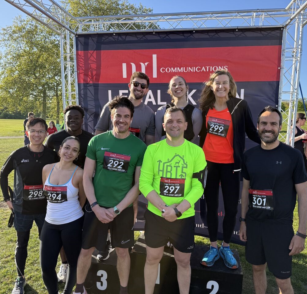 Group of 9 men and women posing for photo in park in front of backdrop that reads MD Communications Legal 5k run. Two women and a man are standing on podium. All wear running clothes and have race numbers pinned to them.